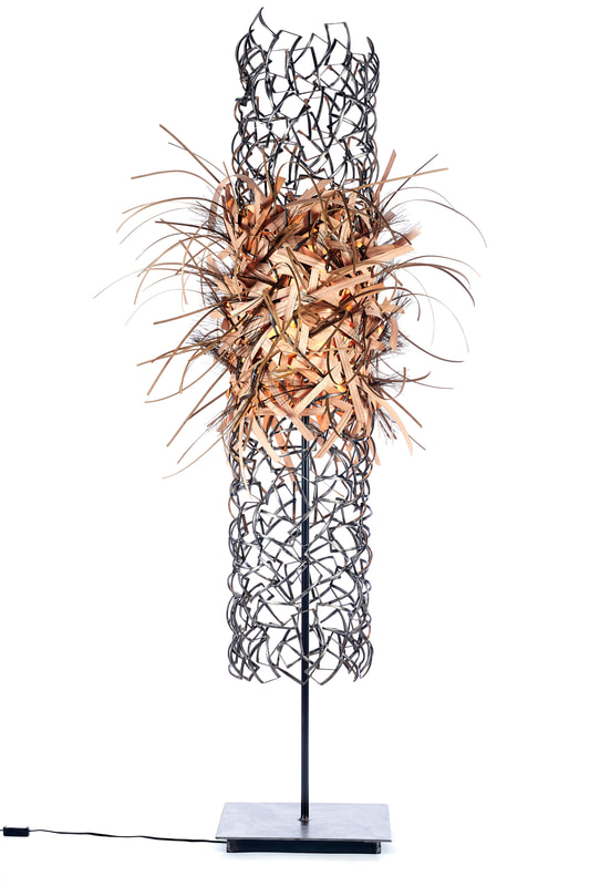Tall sculptural floor light made from scrap metal pieces welded into a long cylinder form from which various colored wooden reed is woven into the center, flaring out in all directions, diffusing the single light bulb. Created by Lucy Slivinski.